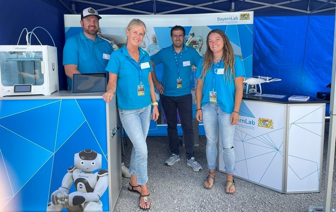 Der Stand vom BayernLab Traunstein auf der BR Radltour auf dem Festgelände in Traunstein. Die vier Mitarbeiter vom BayernLab Traunstein stehen an ihrem Stand auf dem Gelände für die BR Radltour. Es sind 2 Männer und 2 Frauen.Links steht ein 3D-Drucker auf einem Tisch. Die Trennwände im Hintergrund sind in königsblau. Auf einem kleinen weißen Tresen rechts im Bild steht eine weiße Drohne. Die Mitarbeiter tragen alle hellblaue Poloshirts und Jeans.