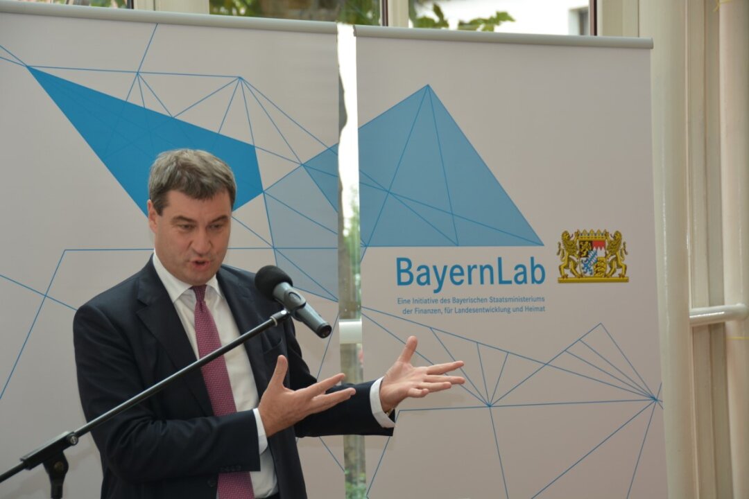 Finanz- und Heimatminister Söder eröffnet das BayernLab Traunstein im Jahre 2016 Finanz- und Heimatminister Söder trägt ein dunkelblaues Jacket, weißes Hemd und Schlips. Er spricht in ein Standmikrofon und hat beide Hände geöffnet. Im Hintergrund ist das Logo vom BayernLab zu sehen und die für die BayernLabs typischen blauen und weißen Polygone.