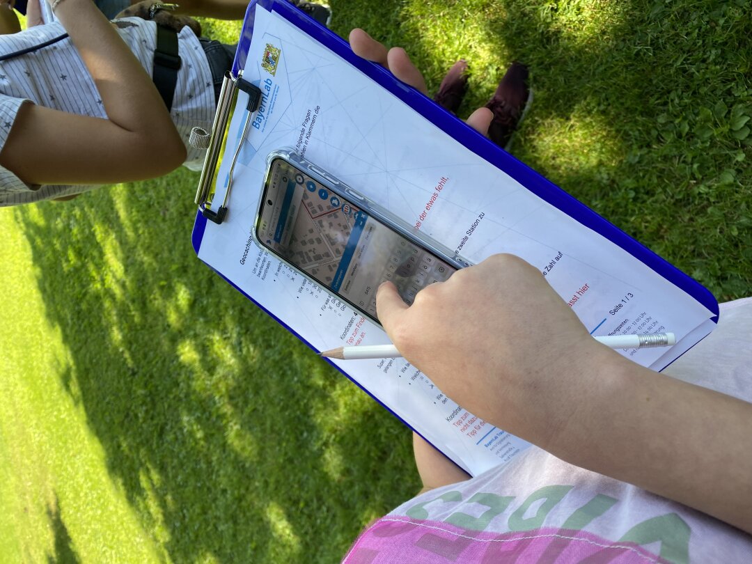 Geocaching: Mit dem BayernAtlas auf digitale Schatzsuche Eine Kinderhand mit einem Bleistift schaut auf ein Clipboard, auf dem ein Blatt Papier eingespannt ist und ein Mobiltelefon mit einem Kartenausschnitt zu sehen ist. Es steht auf einer Wiese.