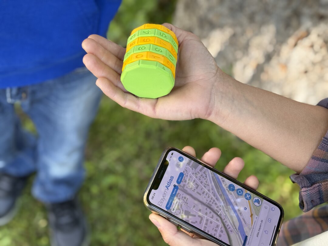 Geocaching: Mit dem BayernAtlas auf Schatzsuche Ein vierstelliges gelbgrünes Zahlenschloss liegt auf einer ausgestreckten Hand, daneben sieht man ein Mobiltelefon mit Geodaten. Im Hintergrund ist verschwommen ein Baum und ein Mensch zu sehen.